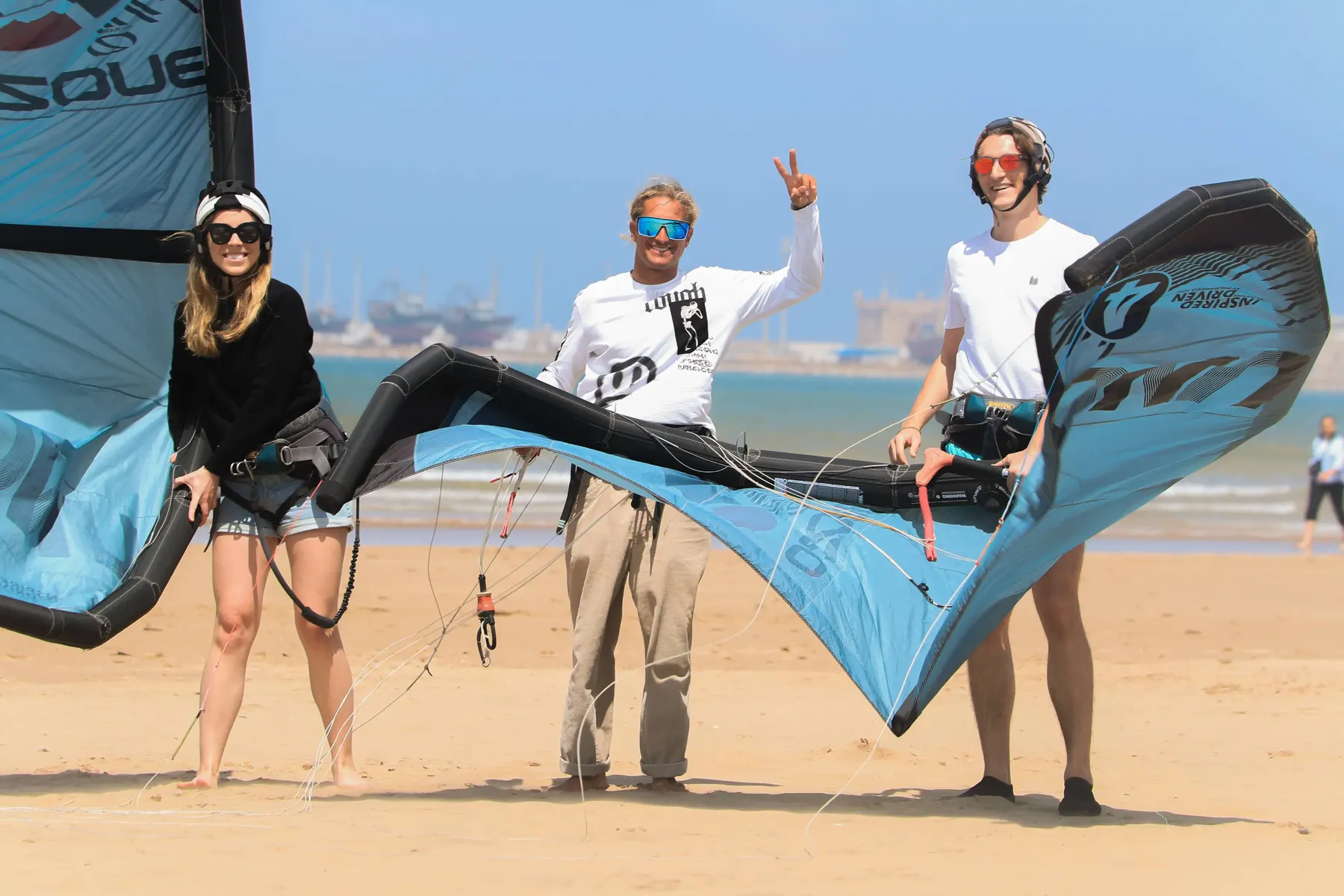 Three people on the beach holding a large blue-and-white kite on the sand, with boats and buildings in the distance