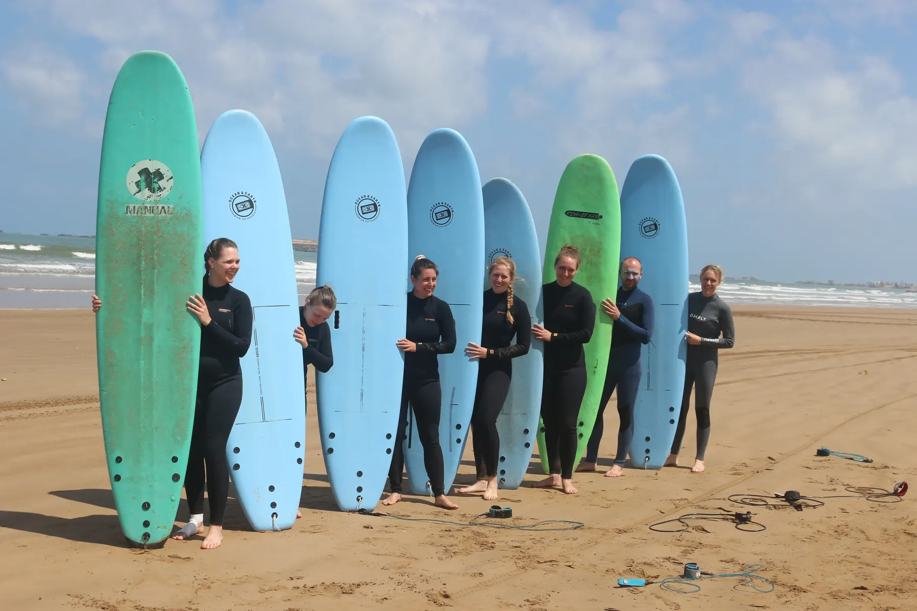 A group of people in wetsuits standing on the sand with tall foam surfboards, the sea behind them