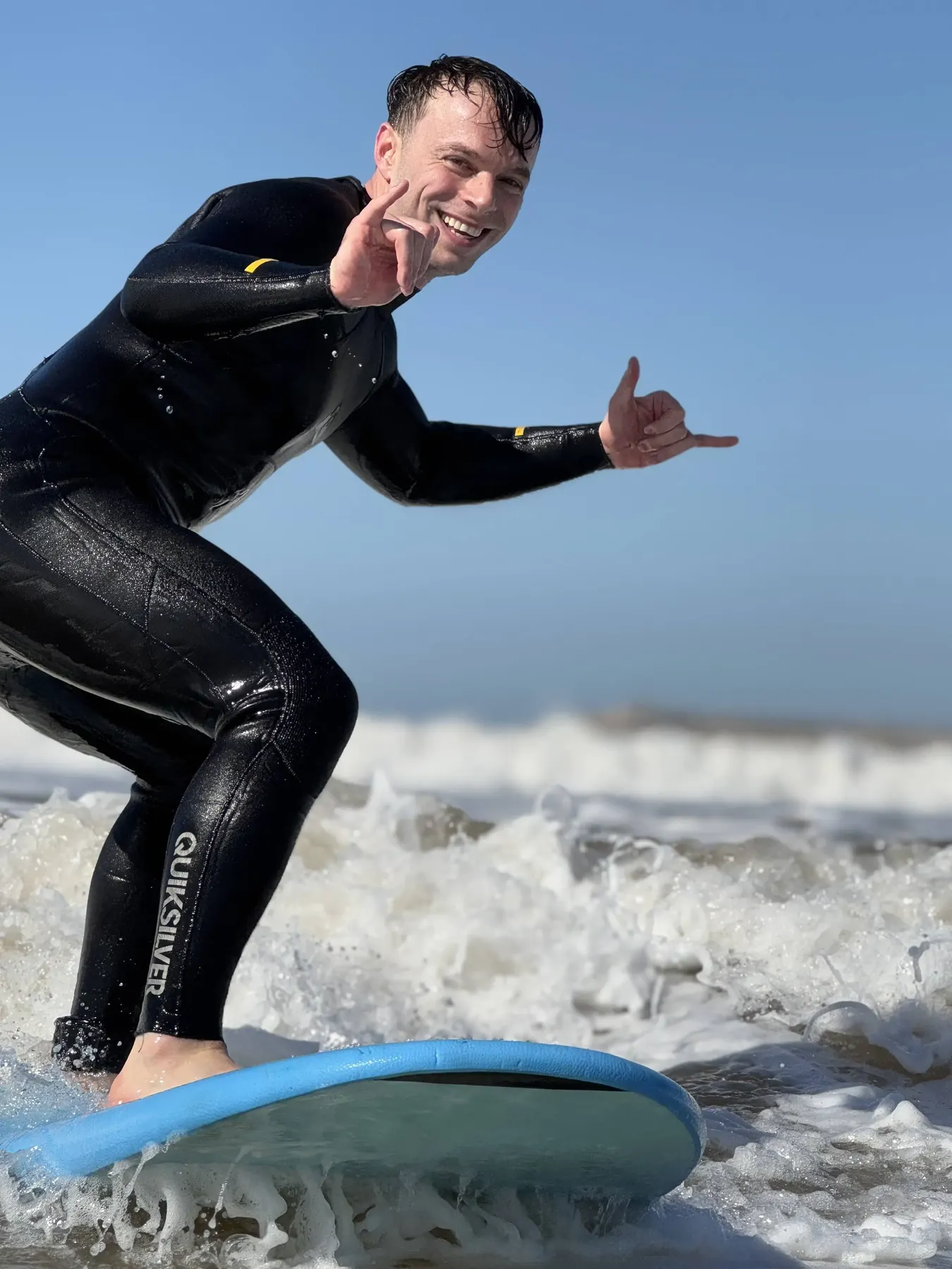 A smiling surfer in a black wetsuit riding white water on a light-blue foam board, making a hand sign