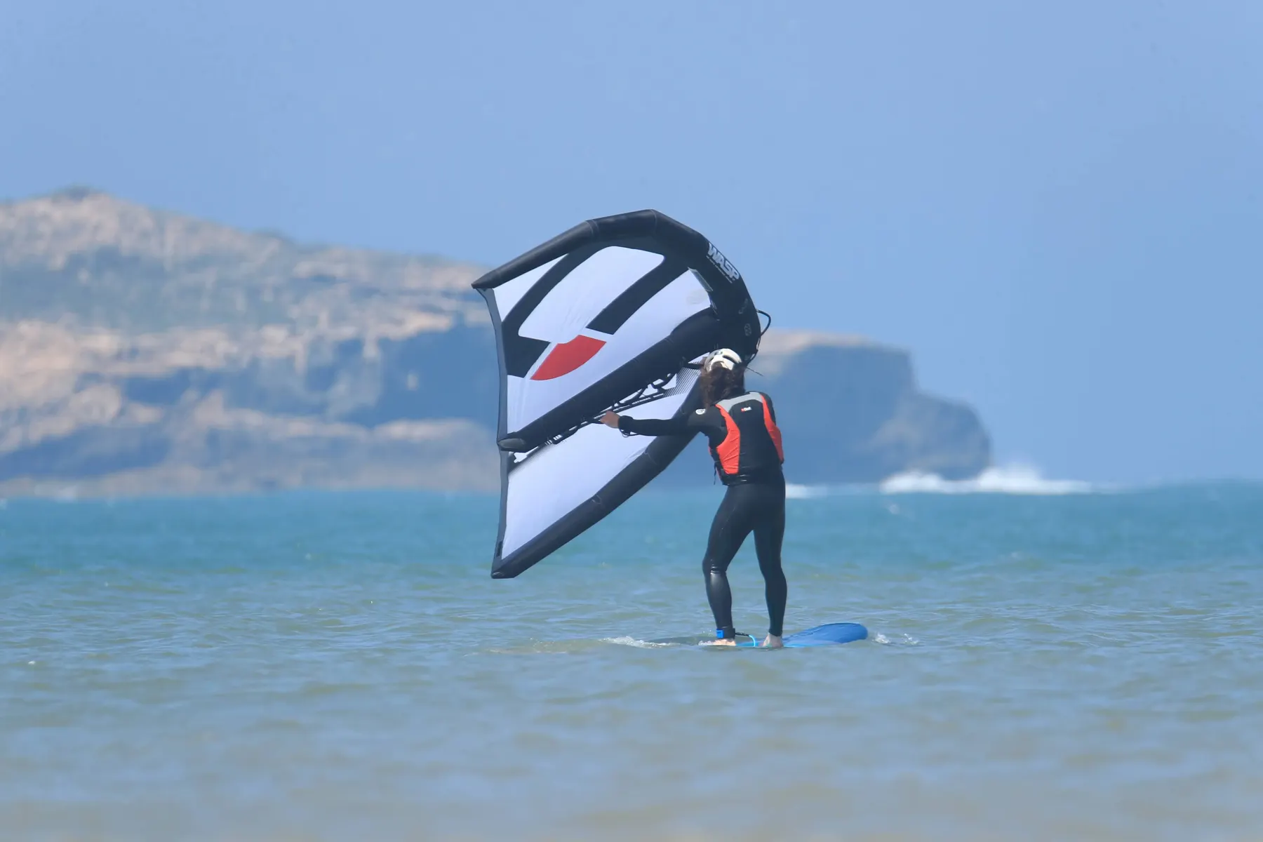 A rider in a wetsuit and orange vest holding a white wing over a board in shallow water, with a headland behind